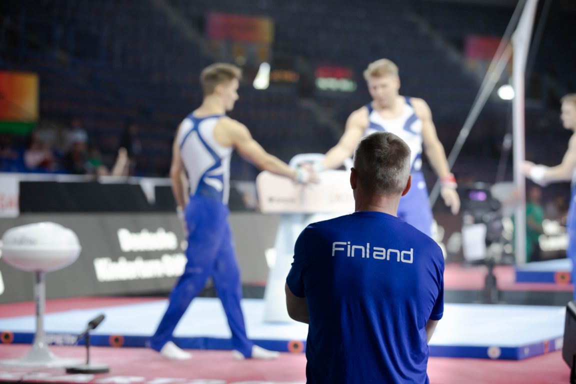 Valmentaja taustallaan kaksi telinevoimistelijamiestä