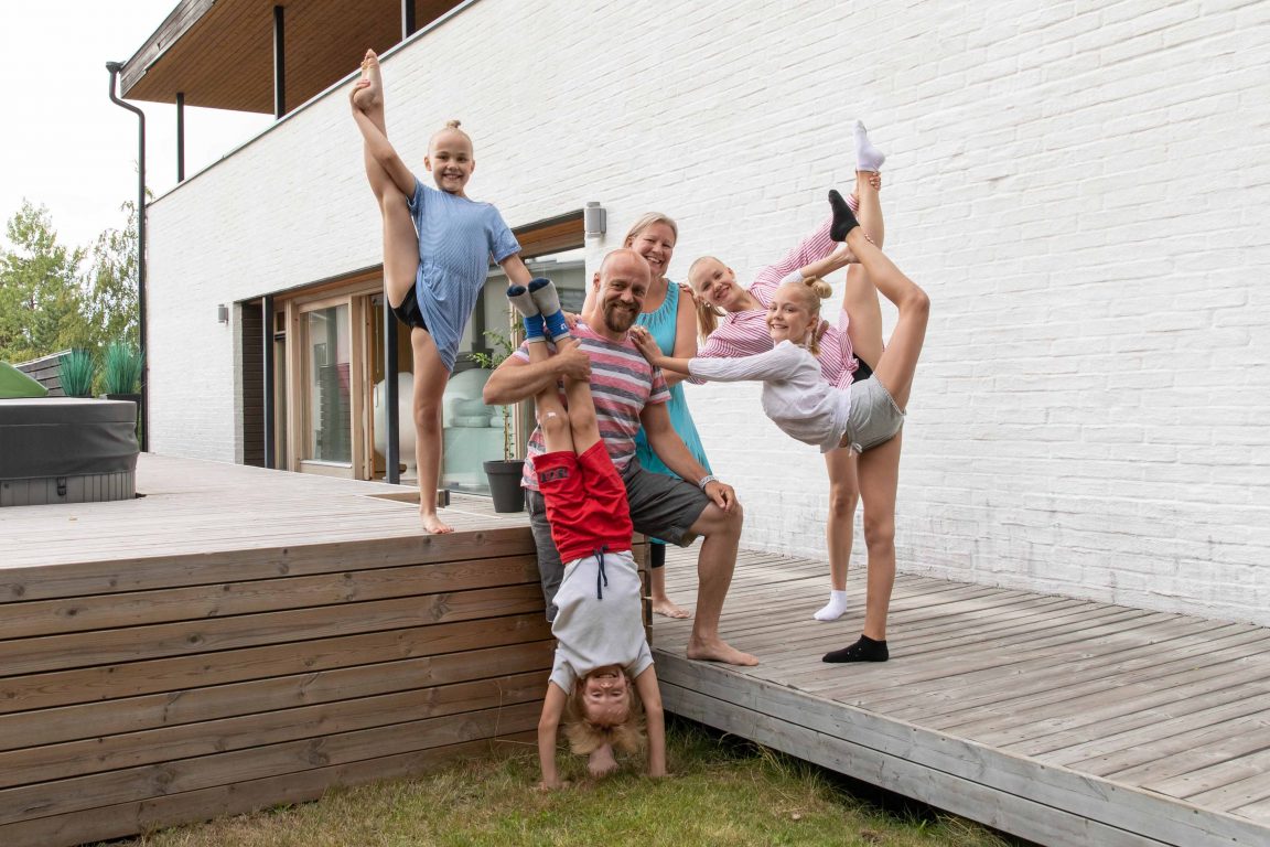 Janne ja perhe poseeraavat kameralla pihalla, lapset voimistelevat käsilläseisonnasta kukkovaakaan.